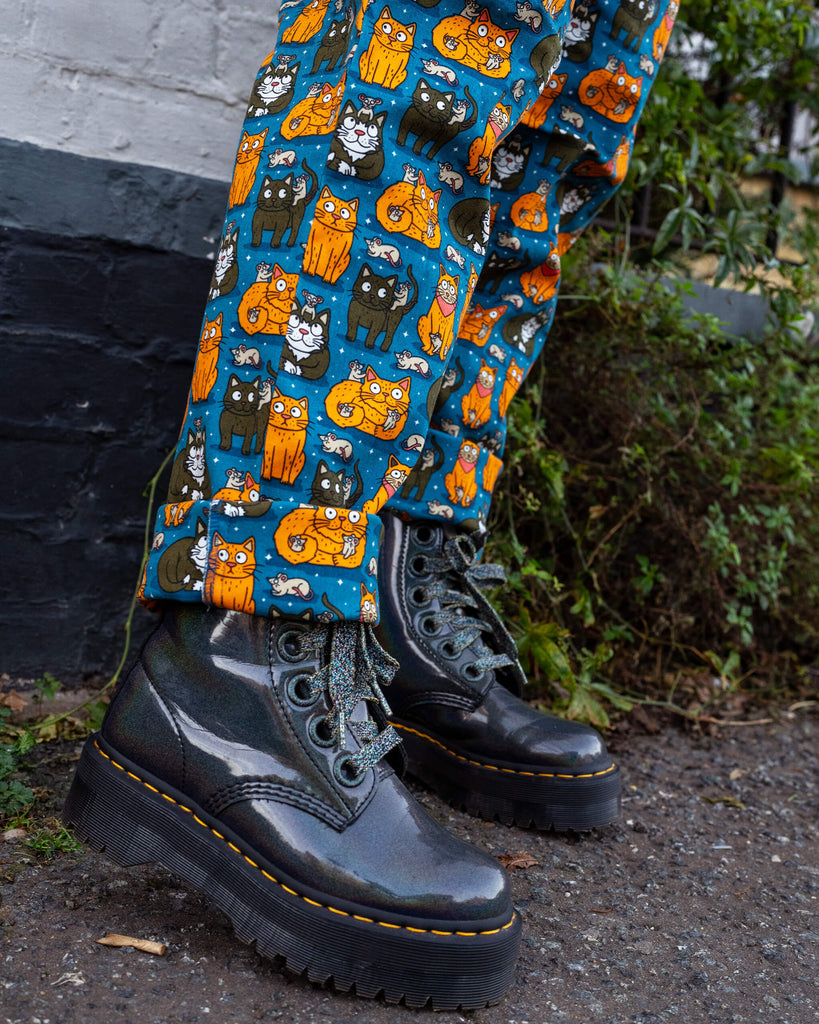 Close-up of legs wearing teal dungarees adorned with an orange and black cat print by Waffle Mama Design, paired with chunky metallic combat boots on a rough pavement background.