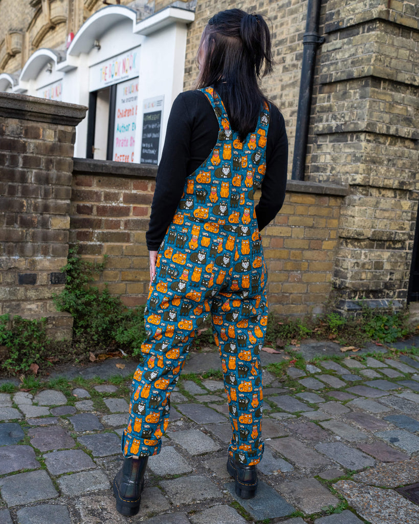 Back view of the model in teal cat and mouse print dungarees, standing in a cobblestone alleyway with brick walls in the background.