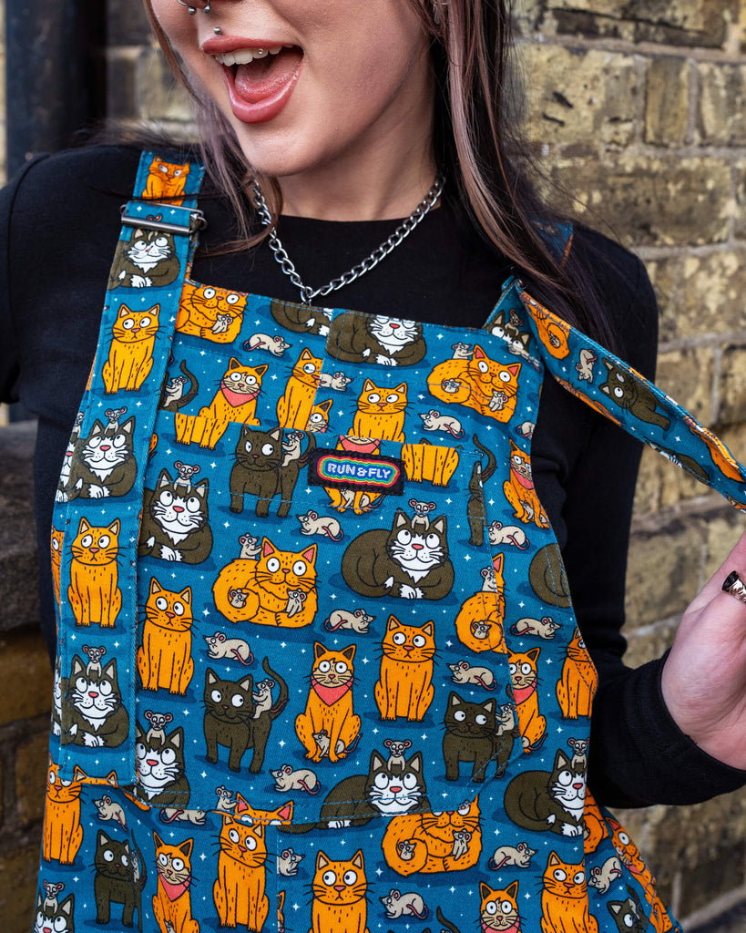 Close-up of model’s upper body, showing the teal dungarees with vibrant orange and black cats, adorned with small grey mice, by Waffle Mama Design, paired with a black top and a chain necklace.