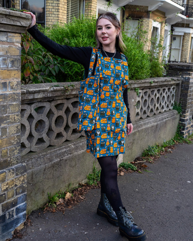 Flo stands in front of a stone railing, smiling, with one hand on their hip, modelling the “Cat and Mouse” print pinafore. The outfit is styled with black tights and black boots for a street-style vibe.