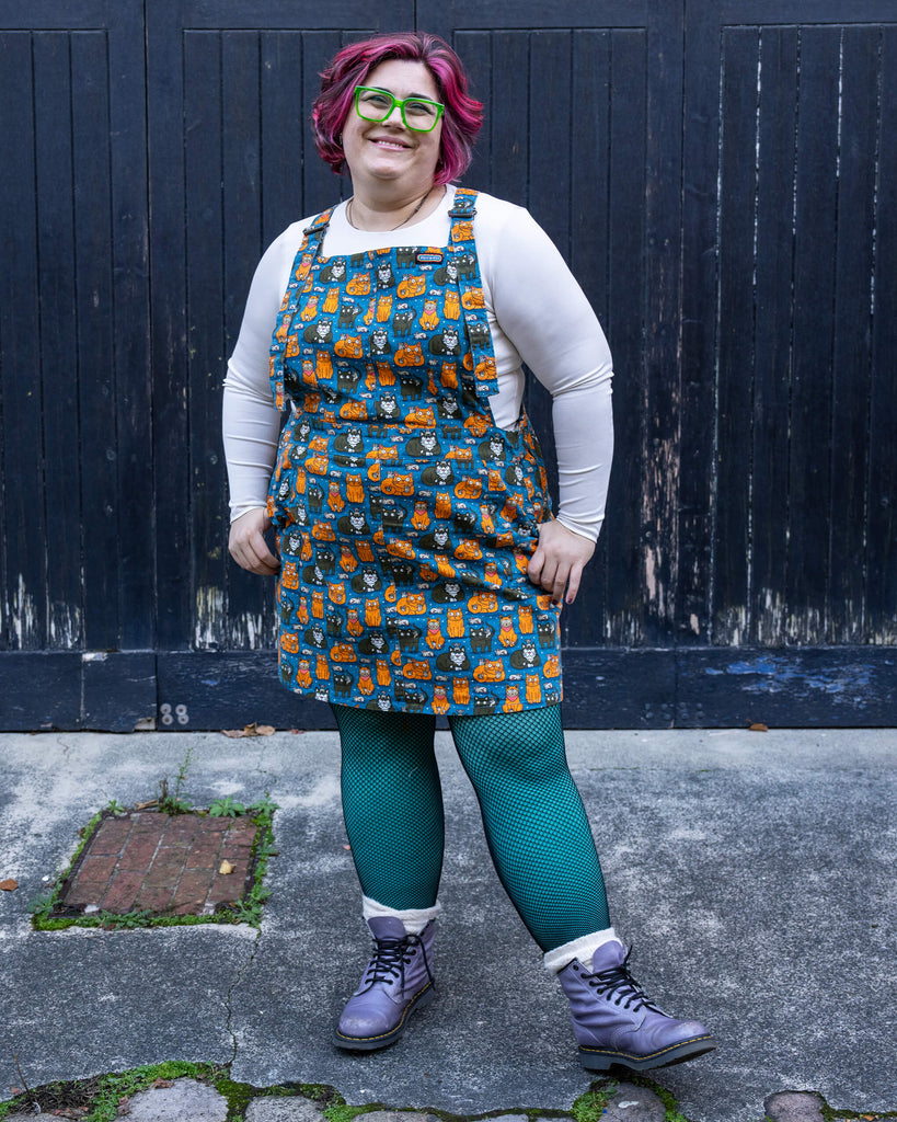 Full-body shot of Jackie in the “Cat and Mouse” pinafore by Run & Fly. The playful design is styled with a cosy white top, colourful tights, and boots, perfect for quirky and fun fashion lovers.