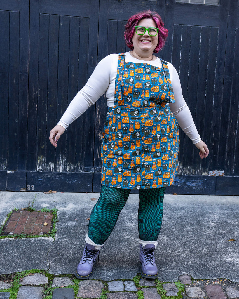 Jackie poses in the Run & Fly “Cat and Mouse” pinafore. The fun and quirky print, filled with orange and black cats alongside tiny mice, creates a cheerful and bold statement look.