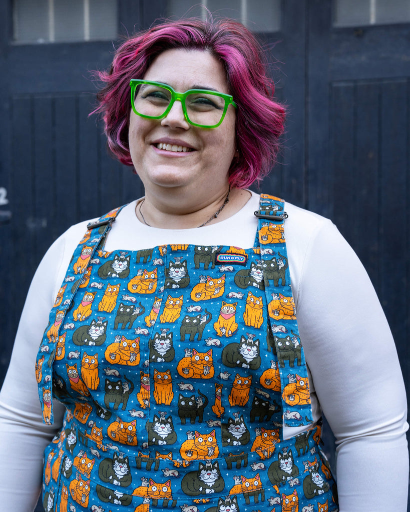Jackie smiles while wearing the “Cat and Mouse” print pinafore by Run & Fly, styled over a white long-sleeve top. The vibrant blue fabric features playful illustrations of cats, mice, and whimsical details, paired with teal leggings and lilac boots.