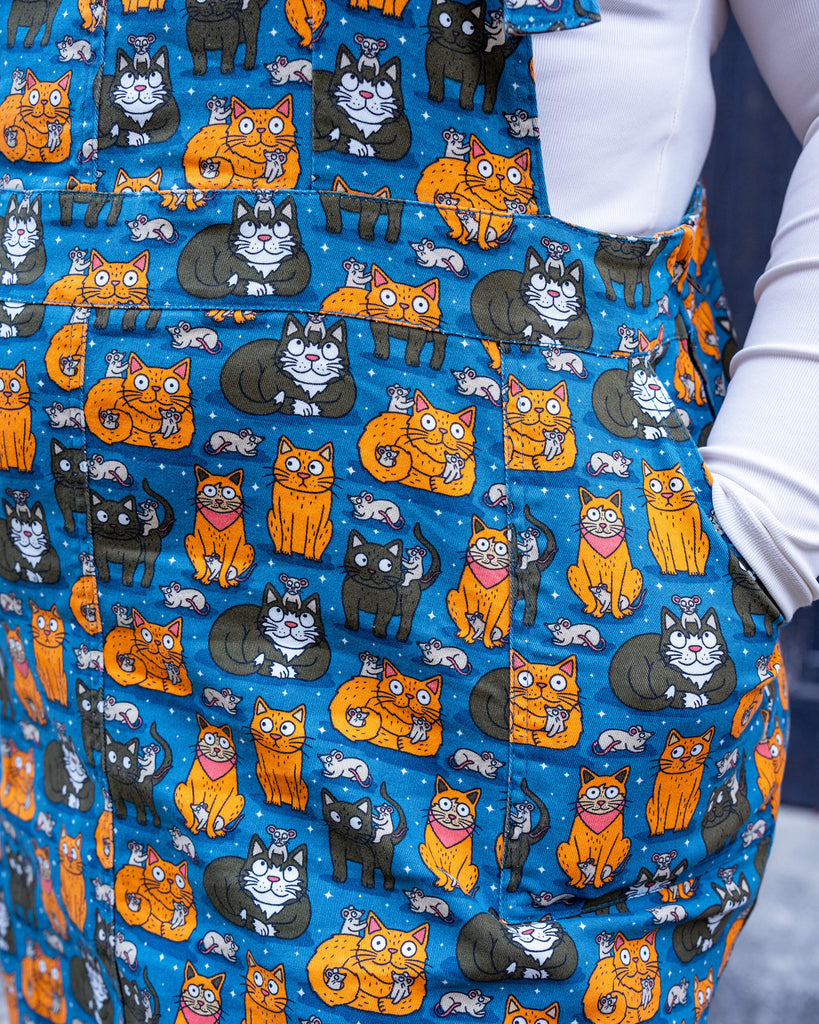 Close-up of the “Cat and Mouse” print pinafore. The design features illustrated cats in various poses with playful mice on a bright blue background, adding charm to this standout piece by Run & Fly.