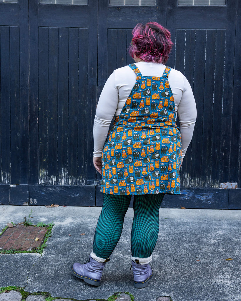 Full-body shot of Jackie in the “Cat and Mouse” pinafore by Run & Fly. The playful design is styled with a cosy white top, colourful tights, and boots, perfect for quirky and fun fashion lovers.