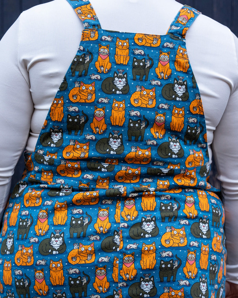  Back view of the “Cat and Mouse” pinafore, showing the seamless continuation of the whimsical print. Jackie pairs it with teal leggings and lilac boots for a playful outfit.