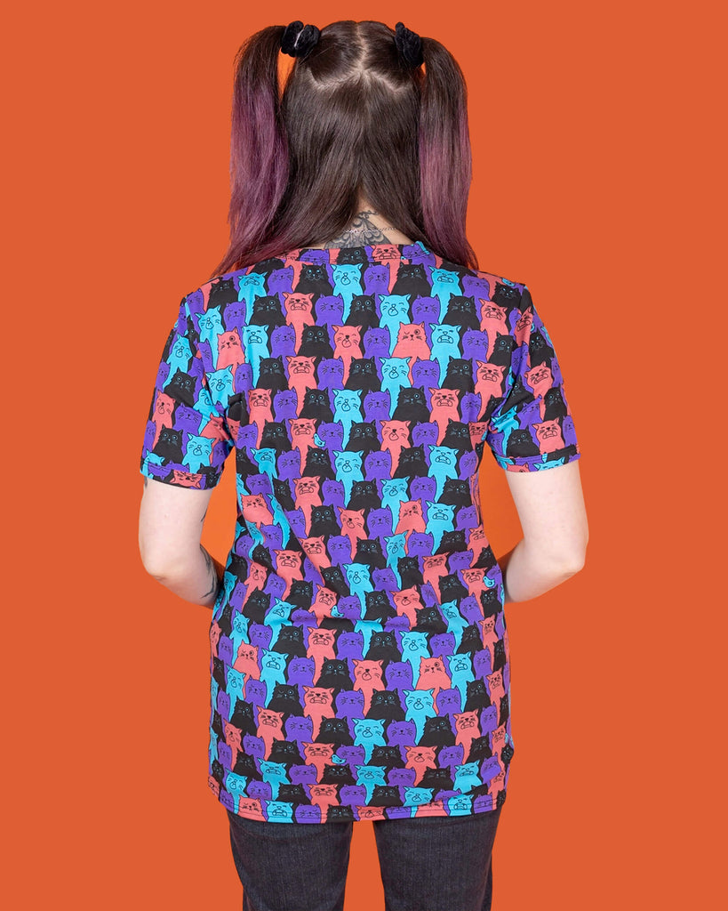 Sarah is stood in front of an orange background wearing the Cat Chorus Short Sleeve Tee with black stonewash denim flares. She is facing forward with one hand flicking her hair and the other hand resting by her side. The cat chorus print is an all over repeating pattern of sky blue, peachy red, purple and black cats pulling various faces and a sporadic sky blue bird.
