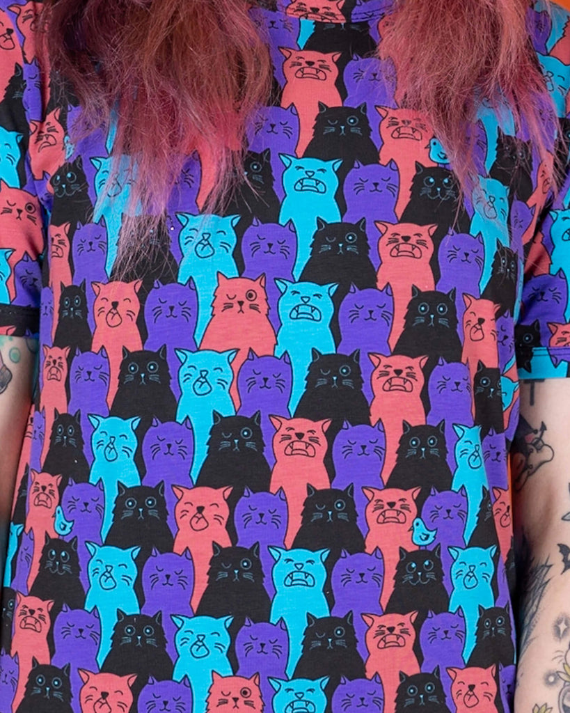 Sarah is stood in front of an orange background wearing the Cat Chorus Short Sleeve Tee with black stonewash denim flares. She is facing forward with one hand flicking her hair and the other hand resting by her side. The cat chorus print is an all over repeating pattern of sky blue, peachy red, purple and black cats pulling various faces and a sporadic sky blue bird.