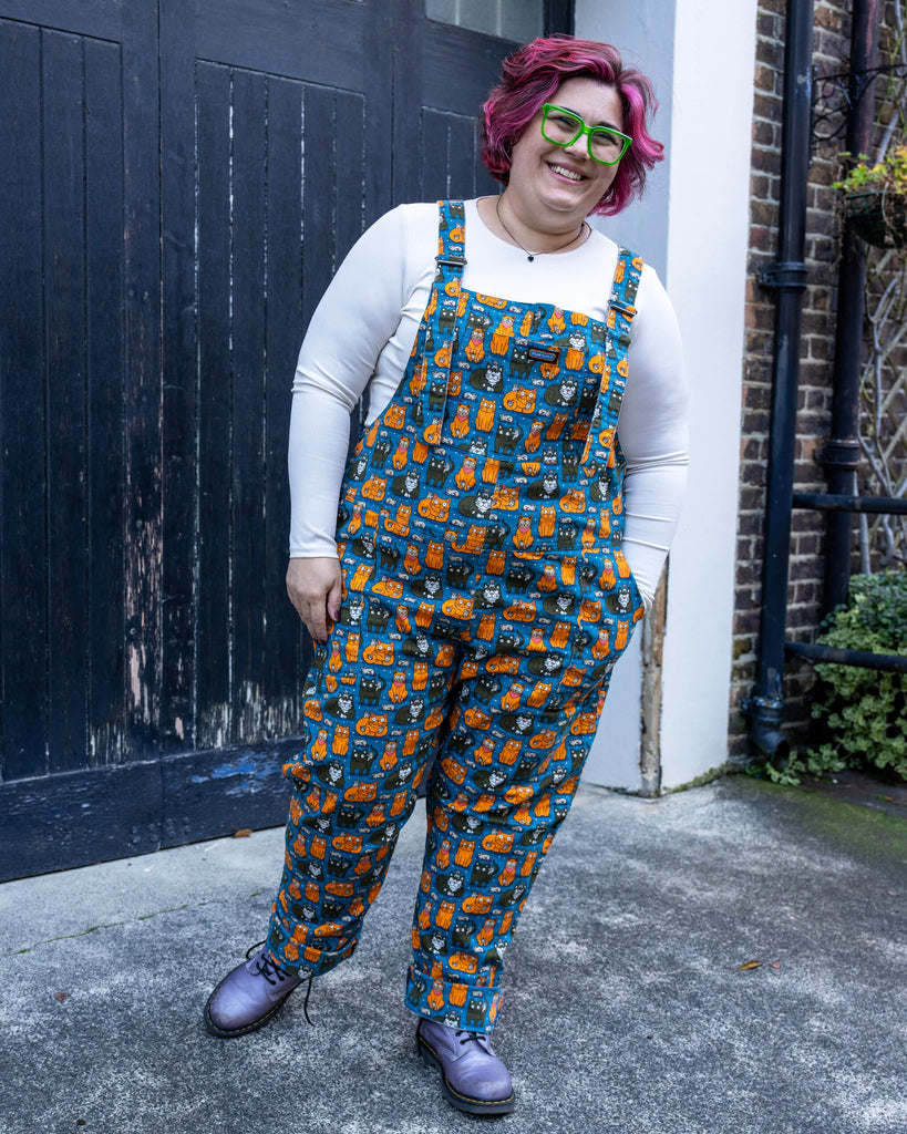 Jackie smiles while wearing the “Cat and Mouse” print dungarees by Run & Fly. The playful design features vibrant blue fabric with quirky illustrations of orange and black cats, tiny mice, and fun details, paired with a white long-sleeve top and lilac boots.