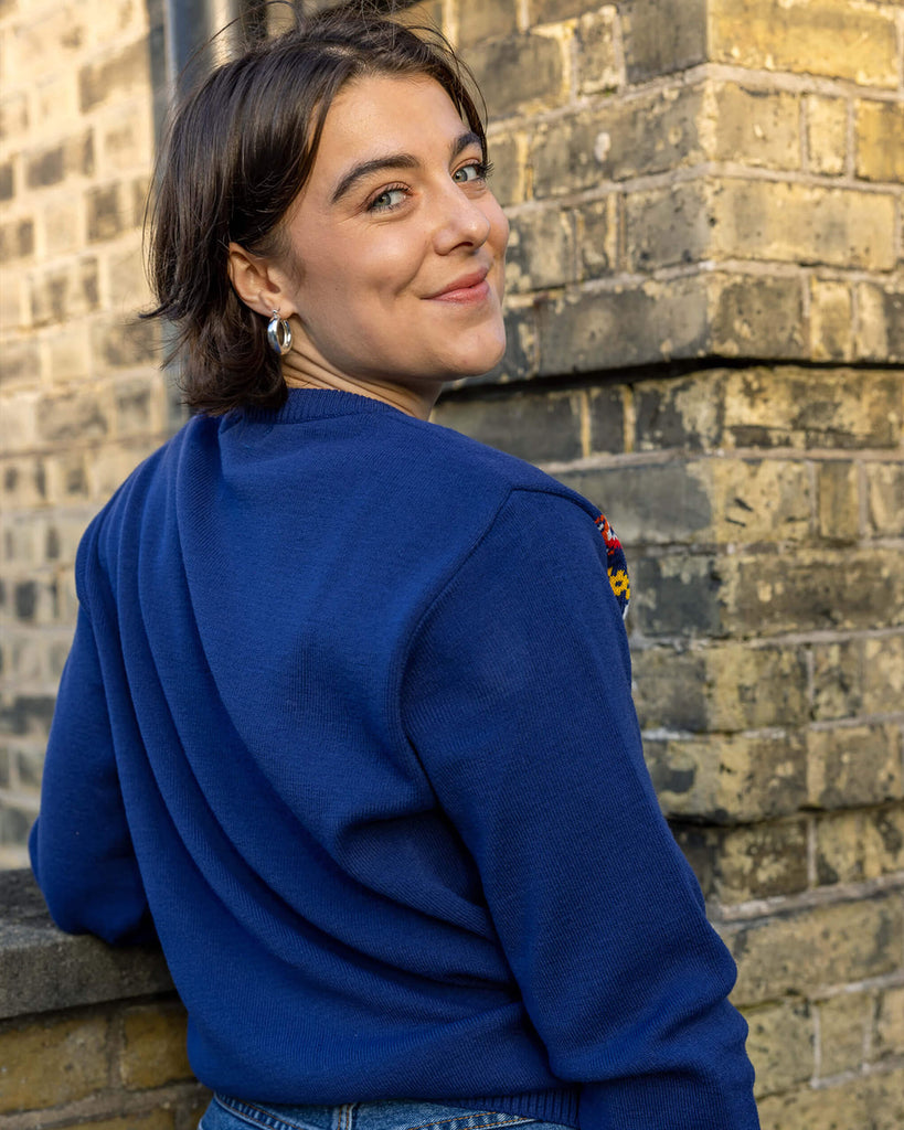 Freya is a white woman in her 20's with short brown hair wearing a blue christmas style jumper with cats on. She is leaning on a brick wall
