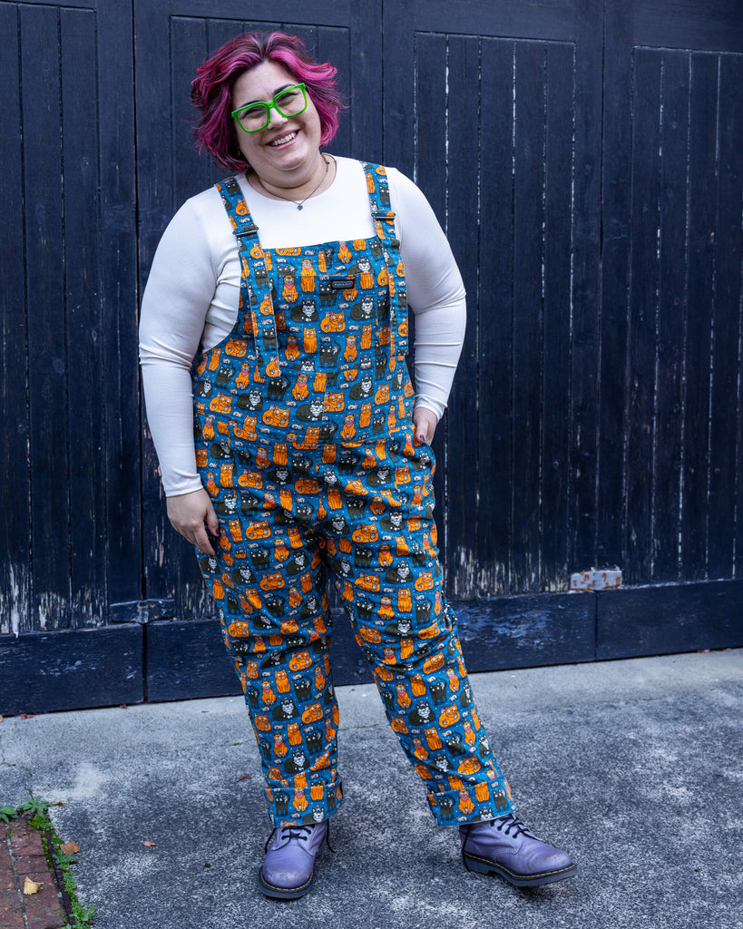 Full-body shot of Jackie wearing the “Cat and Mouse” dungarees by Run & Fly. Styled with teal leggings and cosy boots, the dungarees combine comfort and quirky charm.