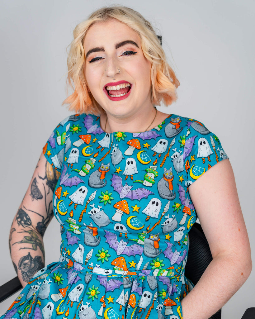 Eliza laughs joyfully while wearing a blue dress featuring the “Cauldron Cats” print by Waffle Mama. The dress is adorned with playful illustrations of cats, ghosts, and mushrooms. Eliza’s tattoos and accessories add to the vibrant, expressive look.