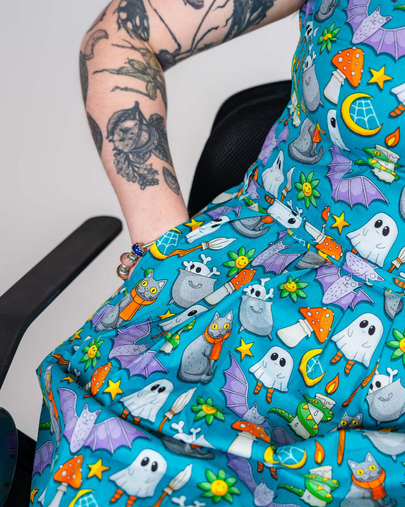 Close-up of Eliza’s tattooed arm and the “Cauldron Cats” dress by Waffle Mama. The print showcases playful Halloween-inspired elements, including ghosts, cauldrons, and bats, against a blue background. The focus is on the detailed artwork and Eliza’s fun, edgy style.