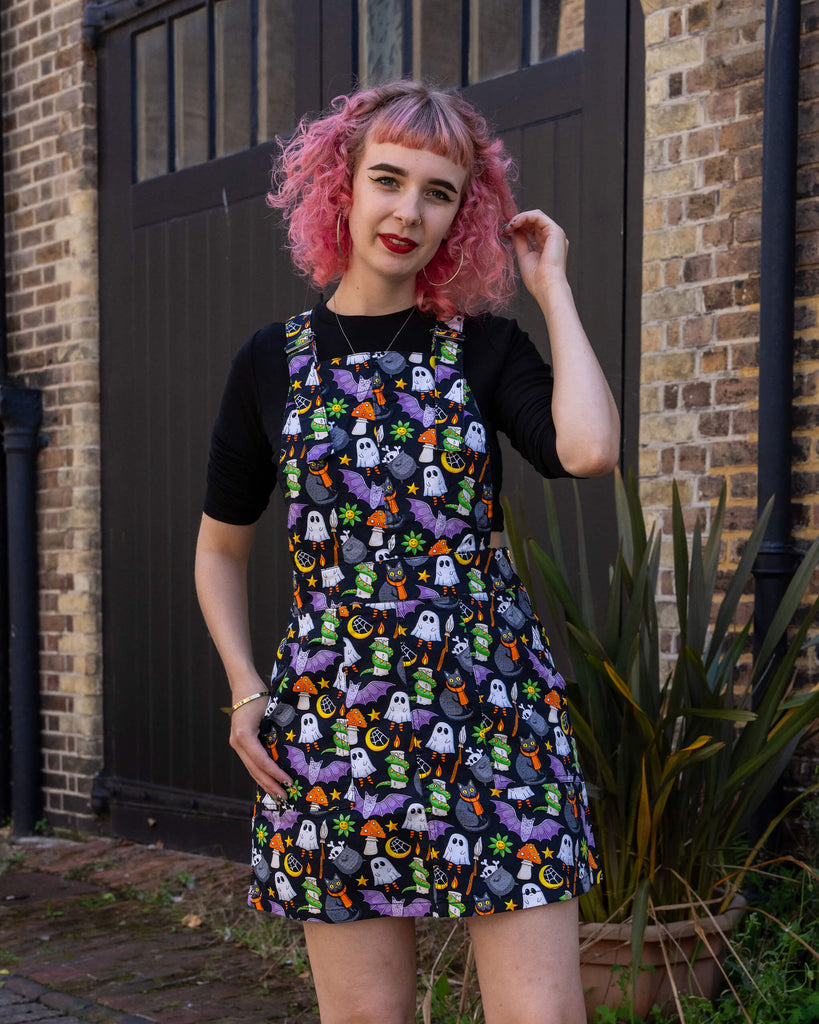A woman with pink hair models a Halloween-themed pinafore dress by Run & Fly, an ethical slow fashion brand. The dress features a fun and vibrant print with ghosts, black cats, bats, and other spooky motifs. She pairs it with a black top and black shoes with red laces, creating a playful, alternative look. Run & Fly is known for its unique, sustainable designs that are perfect for those who appreciate individuality and eco-friendly fashion.