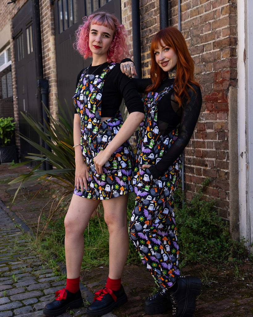 Two women model Halloween-themed outfits by Run & Fly, an ethical slow fashion brand. One wears a playful pinafore dress, and the other sports matching dungarees, both featuring a vibrant print of ghosts, black cats, bats, and other spooky motifs. The outfits are paired with black tops and chunky shoes, creating a fun, alternative style. Run & Fly is celebrated for its unique, sustainable designs that are perfect for those who value individuality and eco-conscious fashion choices.