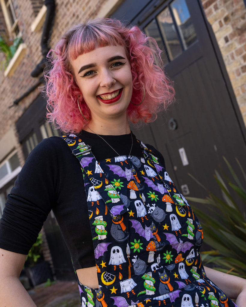 A close-up of a woman with pink hair modelling a Halloween-themed pinafore dress by Run & Fly. The dress features a fun print with ghosts, black cats, bats, and other spooky elements, paired with a black top. Her bright smile and vibrant hair complement the quirky, alternative style. Run & Fly is an ethical slow fashion brand known for its unique, sustainable designs, perfect for those who value individuality and eco-conscious fashion.