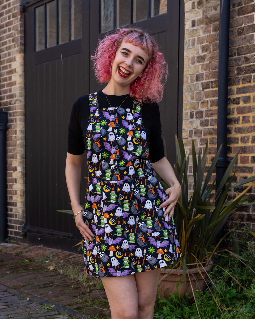 A woman with pink hair models a Halloween-themed pinafore dress by Run & Fly, an ethical slow fashion brand. The dress features a lively print with ghosts, black cats, bats, and other spooky elements, creating a fun, quirky look. She pairs the dress with a black top and black shoes with red laces, adding a bold contrast to her outfit. Run & Fly is renowned for its sustainable, unique designs that cater to those who value individuality and eco-conscious fashion.