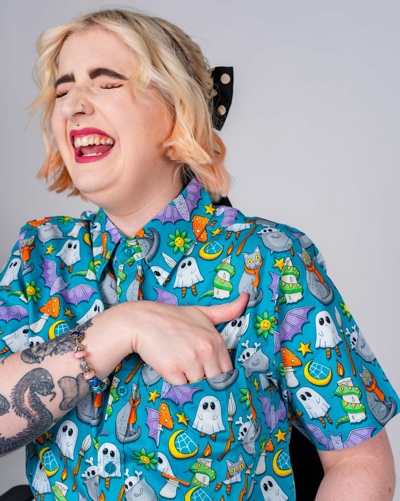 A person with blonde hair and pink tips, laughing while wearing a bright blue Run & Fly shirt covered in playful Halloween-themed designs including ghosts, cats, and cauldrons. Ethical slow fashion brand.