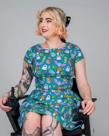 Eliza, a non-binary person in a wheelchair, models a blue dress with a colourful “Cauldron Cats” print by Waffle Mama. The whimsical design features ghosts, cats, mushrooms, and stars. Eliza’s blonde hair is styled with a black polka dot bow, and they are smiling while showcasing the fun, playful design.