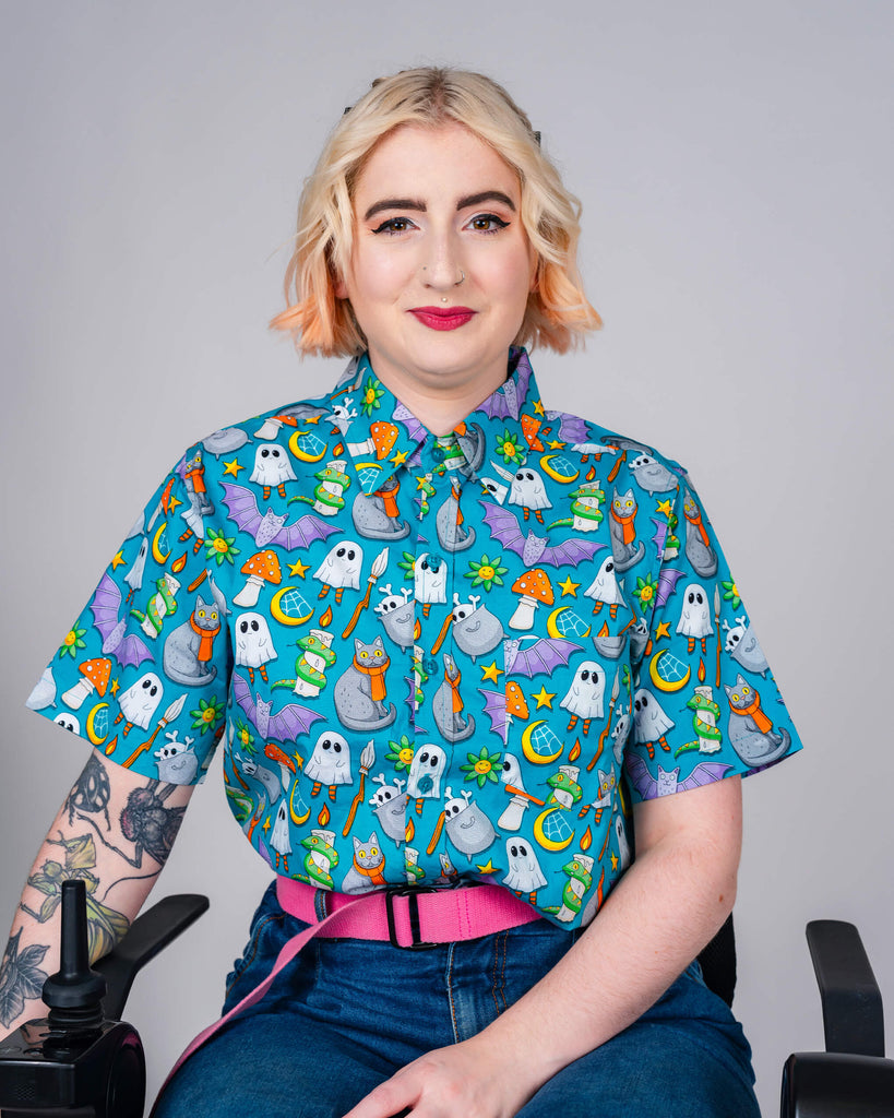 A person sits smiling in a vibrant blue Run & Fly shirt covered in fun, spooky-themed illustrations like ghosts, cauldrons, and cats. Ethical slow fashion from a UK brand.