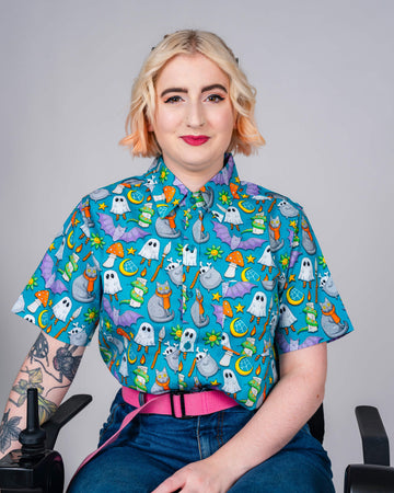 A person sits smiling in a vibrant blue Run & Fly shirt covered in fun, spooky-themed illustrations like ghosts, cauldrons, and cats. Ethical slow fashion from a UK brand.