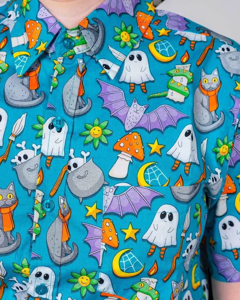 A close-up of a colourful Run & Fly shirt showcasing whimsical Halloween designs with friendly ghosts, cats, and cauldrons on a vibrant blue background. Ethical UK slow fashion.