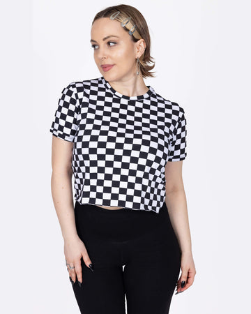 The Checkerboard Short Sleeve Crop Tee worn by a femme model with short blonde hair and high rise black leggings in front of a white studio background. She is facing forward smiling to camera with both hands resting on her hips. The crop tee has an all over black and white checkerboard print with a raw hem for a y2k 90s style.