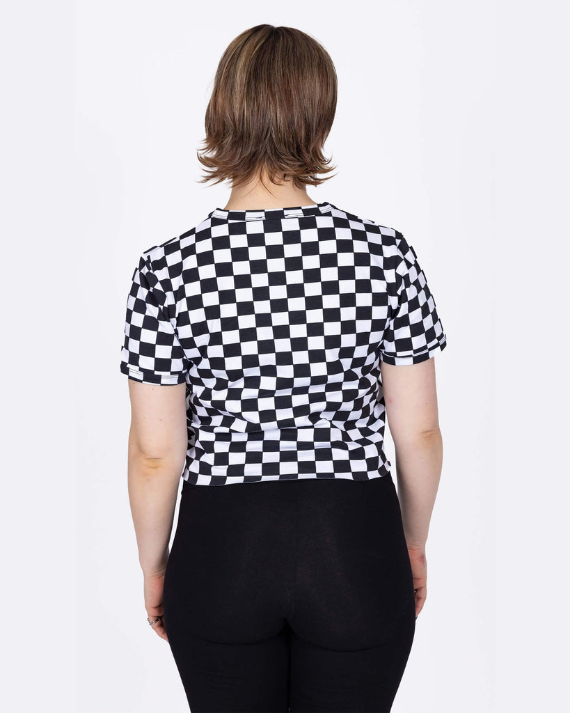 The Checkerboard Short Sleeve Crop Tee worn by a femme model with short blonde hair and high rise black leggings in front of a white studio background. She is facing away from camera with both hands resting by her side. The crop tee has an all over black and white checkerboard print with a raw hem for a y2k 90s style.