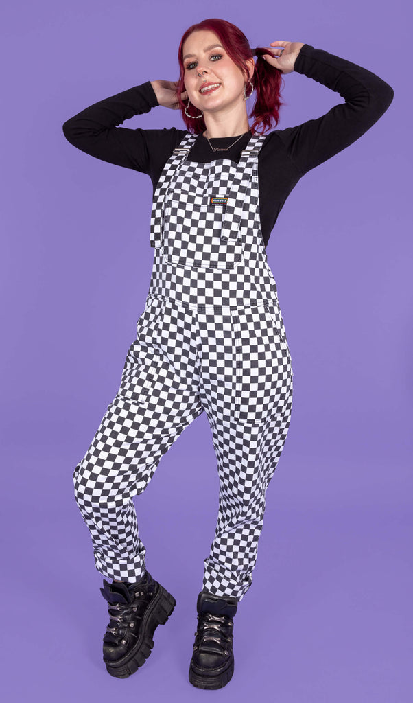 Florence, a femme model with red hair and piercings, is stood in a photography studio on hove in front of a purple backdrop wearing Washed Black & White Checkerboard Stretch Twill Dungarees with a long sleeved black top underneath and chunky black shoes. Florence is smiling at the camera with one leg bent and her hands in her hair.