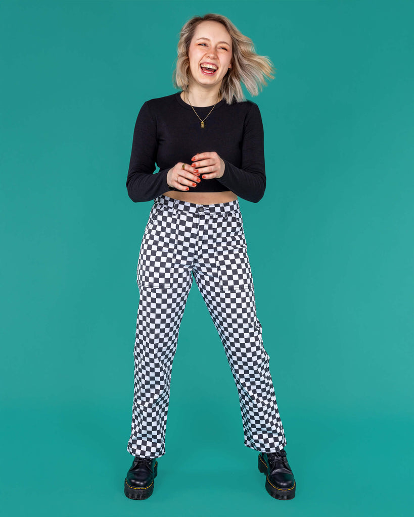 Amy a blonde white model with short hair is smiling and laughing wearing a black long sleeved top and black and white checkerboard trousers with black boots on a green background
