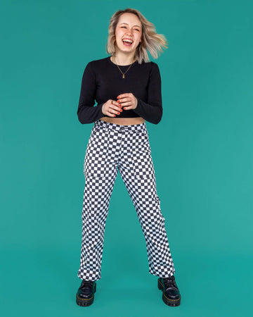 Amy a blonde white model with short hair is smiling and laughing wearing a black long sleeved top and black and white checkerboard trousers with black boots on a green background