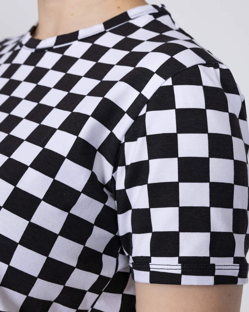 Close up of the Checkerboard Short Sleeve Crop Tee worn by a femme model with short blonde hair in front of a white studio background. She is facing left smiling to with both arms resting by her side to highlight the sleeve stitching. The crop tee has an all over black and white checkerboard print with a raw hem for a y2k 90s style.