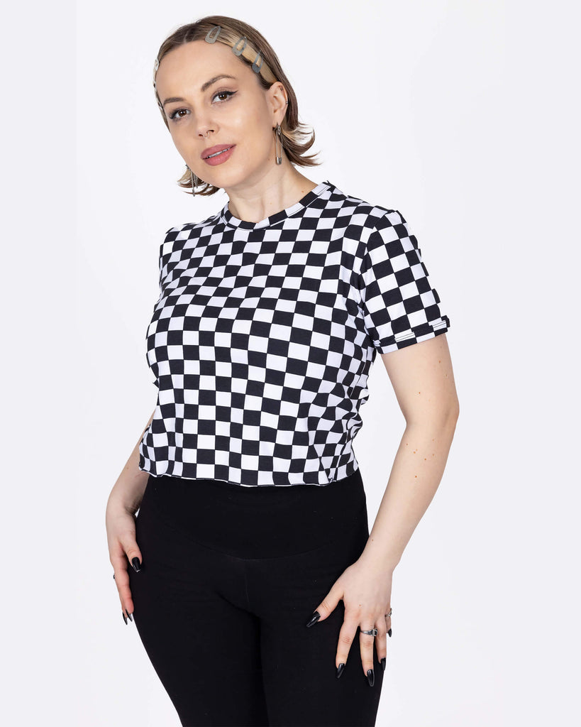 The Checkerboard Short Sleeve Crop Tee worn by a femme model with short blonde hair and high rise black leggings in front of a white studio background. She is facing forward smiling to camera with both hands resting on her hips. The crop tee has an all over black and white checkerboard print with a raw hem for a y2k 90s style.