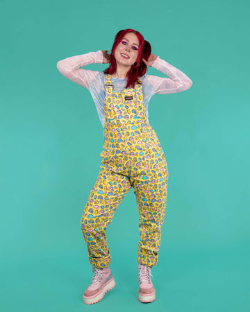 Run & Fly x Sugar & Sloth Chonky Dino Stretch Corduroy Dungarees worn by Flo on a teal background. The dungarees are a soft yellow base colour with various chonky kawaii dinosaurs in pastel blue blue, yellow and pink with rainbow star bursts. Flo has short red hair in bunches, neon pink eye makeup and tattoos, she has styled the dungarees with a pastel pink and blue mesh long sleeve top and pastel pink and blue platform boots. She is facing forward with both arms holding her hair whilst smiling to camera.