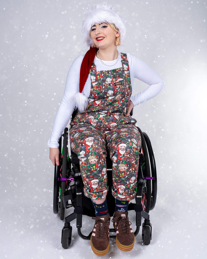 Eliza, a non-binary disabled person using a wheelchair, smiles while wearing Christmas-themed dungarees featuring festive characters, including Santa, elves, and snowmen, by Lauren Ash Design for Run & Fly. They pair the outfit with a white long-sleeve shirt and a fluffy Santa hat, adding a cheerful holiday vibe.
