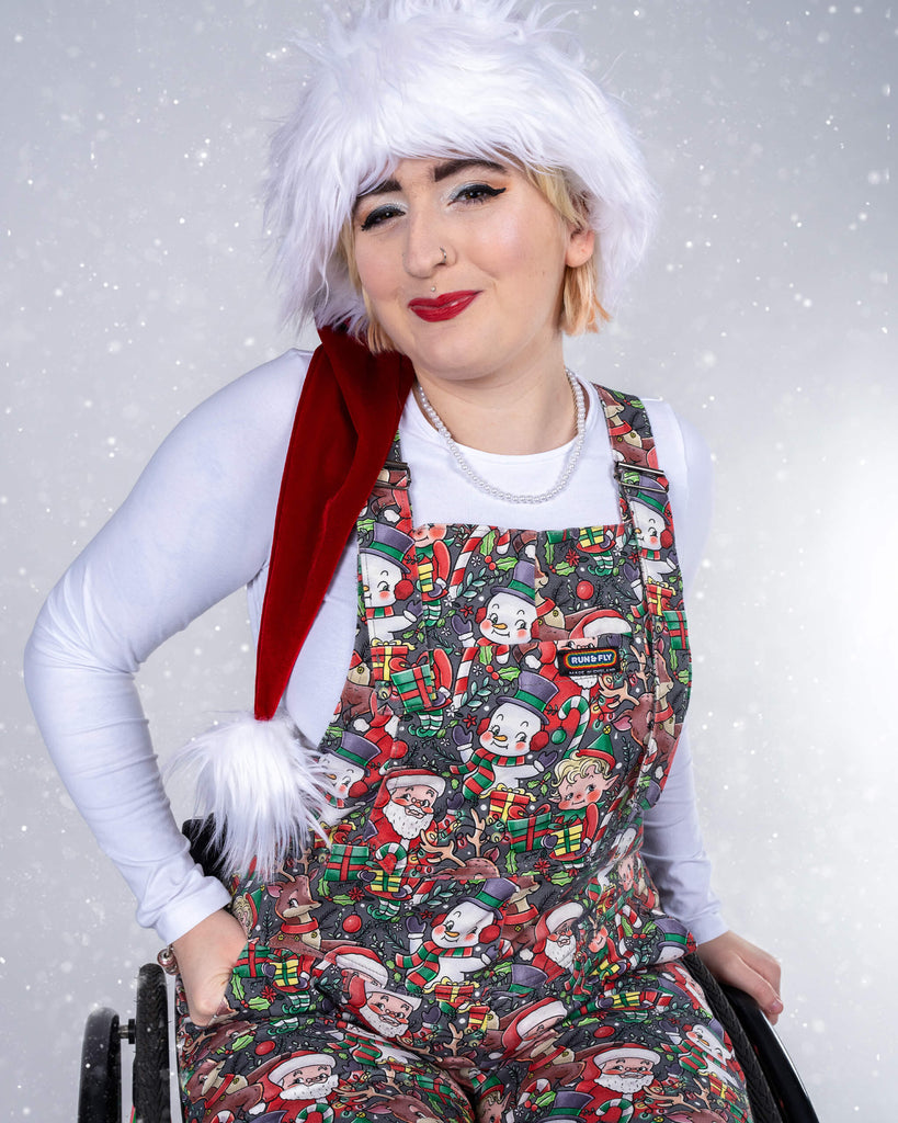Eliza, in their wheelchair, strikes a joyful pose, tilting slightly as they smile and look off to the side. The Christmas-themed dungarees, featuring a detailed holiday print, create a cheerful holiday look when paired with a Santa hat and white shirt.