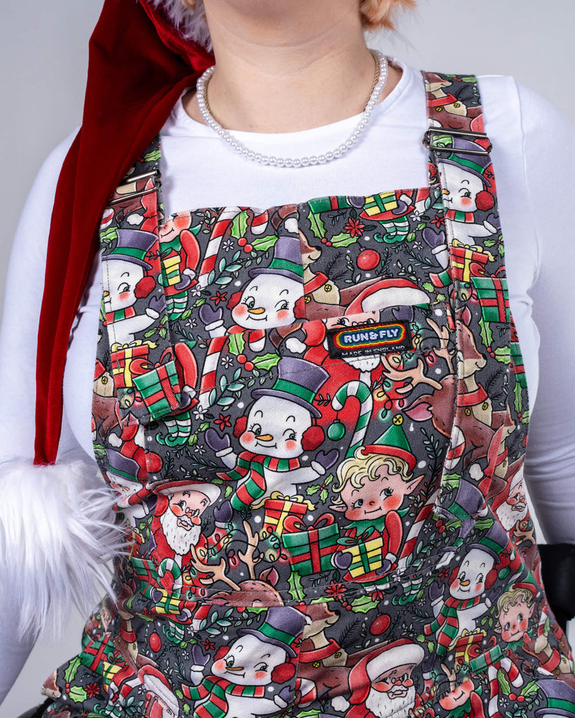 Close-up of the chest area of Eliza’s Christmas-themed dungarees by Lauren Ash Design for Run & Fly. The print is filled with charming holiday characters like Santa and elves, accented by bright festive colours. Eliza’s pearl necklace and Santa hat complement the seasonal attire.