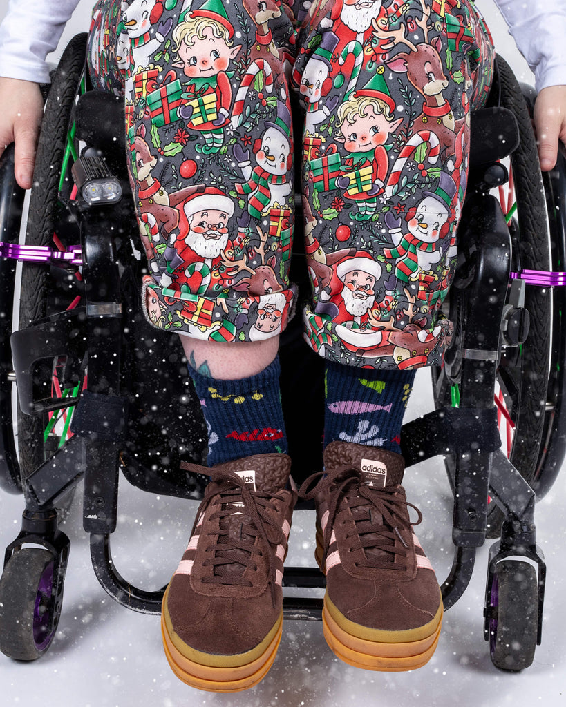  A closer view of Eliza’s legs, highlighting the playful Christmas print on their rolled-up dungaree trousers. The design features holiday characters and vibrant colours. Eliza’s brown sneakers and festive socks complete this cheerful holiday look.