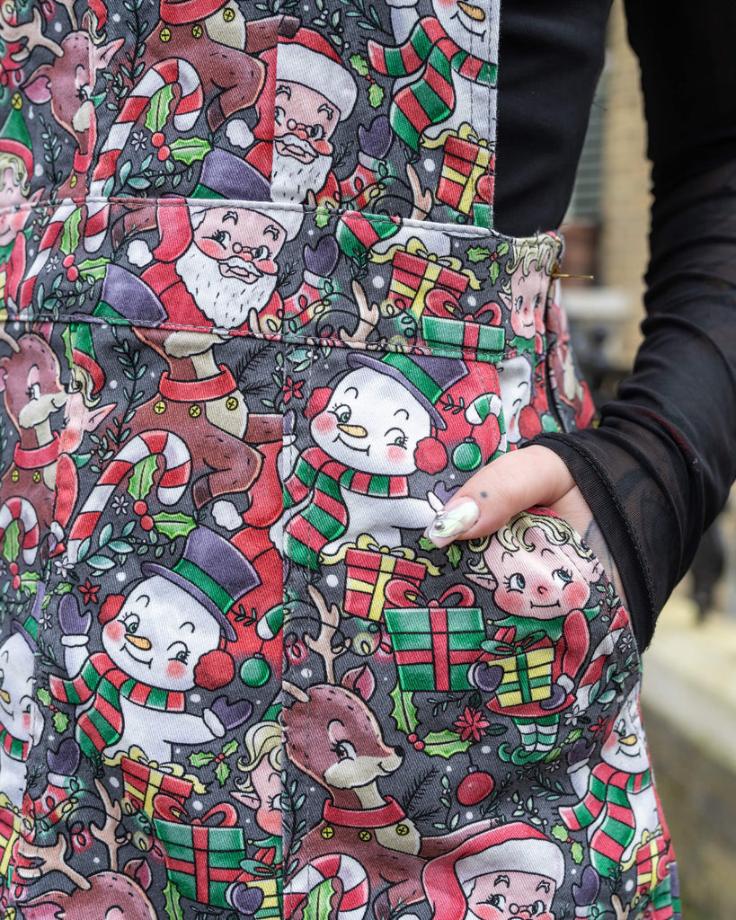 Close-up of Run & Fly's Christmas-themed pinafore pocket, showcasing intricate designs of Santa, snowmen, reindeer, and presents on a dark background. The ethical slow fashion brand highlights playful and vibrant festive aesthetics.