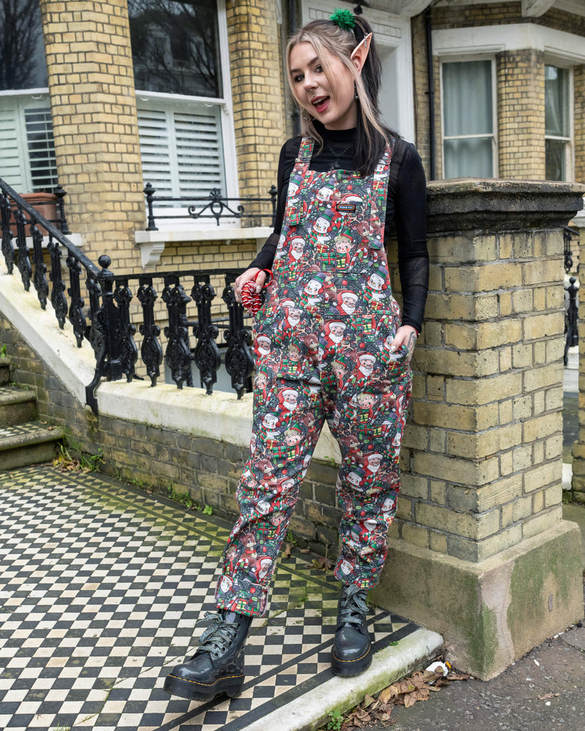 Front view of a model in Run & Fly Christmas-themed dungarees with playful festive designs. Styled with elf-inspired accessories, green hair ties, and chunky black boots, the outfit radiates holiday fun. Captured in a vintage outdoor setting.