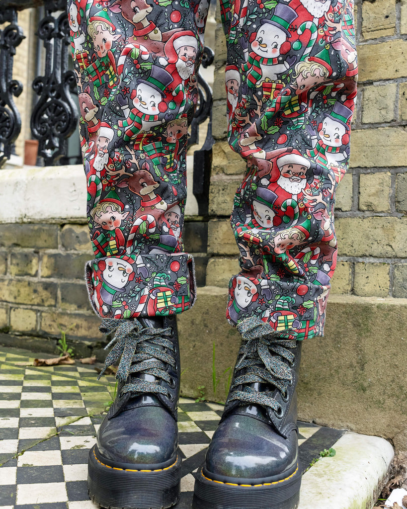 Close-up of Run & Fly Christmas-themed dungarees showing detailed festive prints of Santa, reindeer, and snowmen. The model styles them with rolled-up cuffs and glittery black boots, showcasing the fun and ethical slow fashion design.