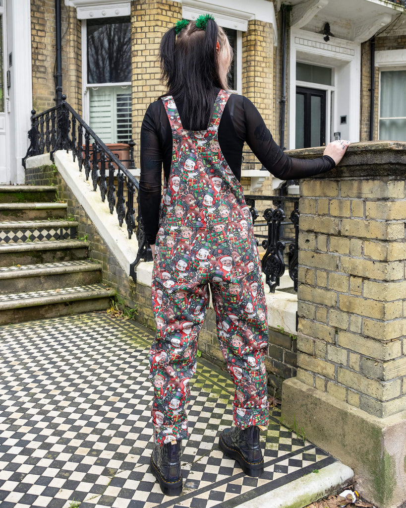 Back view of a model wearing Run & Fly Christmas-themed dungarees with festive illustrations of Santa, elves, snowmen, and reindeer. Paired with black mesh sleeves and chunky black boots, the look adds a quirky holiday vibe. Photographed outdoors on a vintage chequered path with a brick house backdrop.