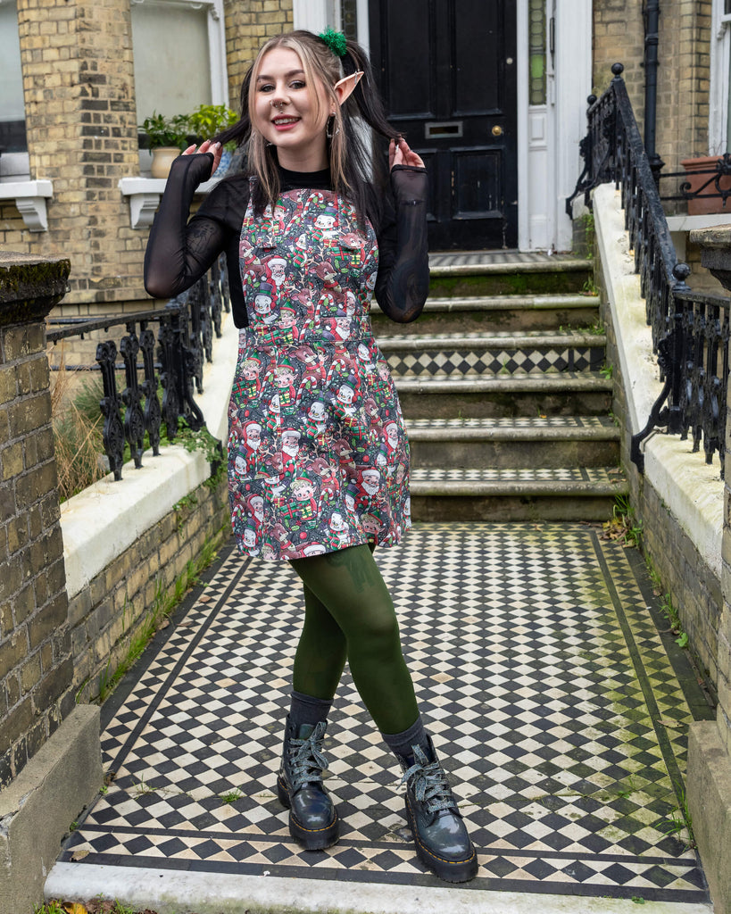 Model striking a playful pose in a Run & Fly Christmas-themed pinafore with festive prints of Santa, snowmen, and reindeer. Styled with green tights, black boots, and elf ear accessories, this outfit brings cheerful vibes, photographed against a historic brick building.
