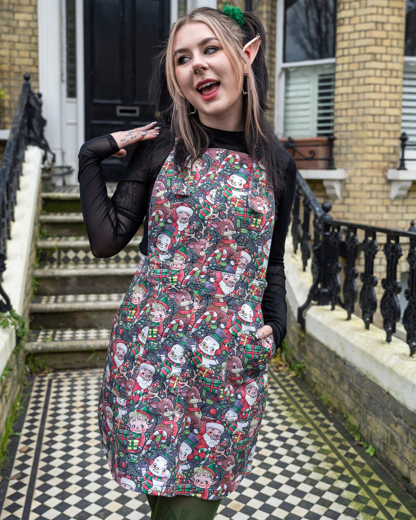 Front view of a model in a Run & Fly Christmas-themed pinafore dress, featuring Santa, elves, and reindeer. Styled with black mesh sleeves, green tights, and elf ear accessories, the look combines festive spirit and quirky fashion. Shot on a vintage-style chequered pathway.