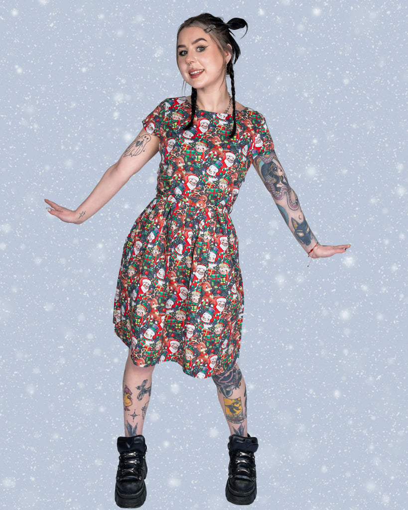 Flo strikes a joyful pose in the Christmas Cuties tea dress, showing off the festive print filled with Santas, elves, reindeer, and presents. Her black boots and braided hairstyle add a modern edge to the wintery snowflake backdrop.