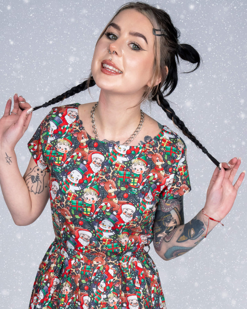 Flo poses playfully, holding her braided pigtails, wearing a tea dress featuring the Christmas Cuties print by Lauren Ash Design. The design shows festive characters like Santa, reindeer, and snowmen with colourful holiday decorations. A snowy background completes the whimsical scene.