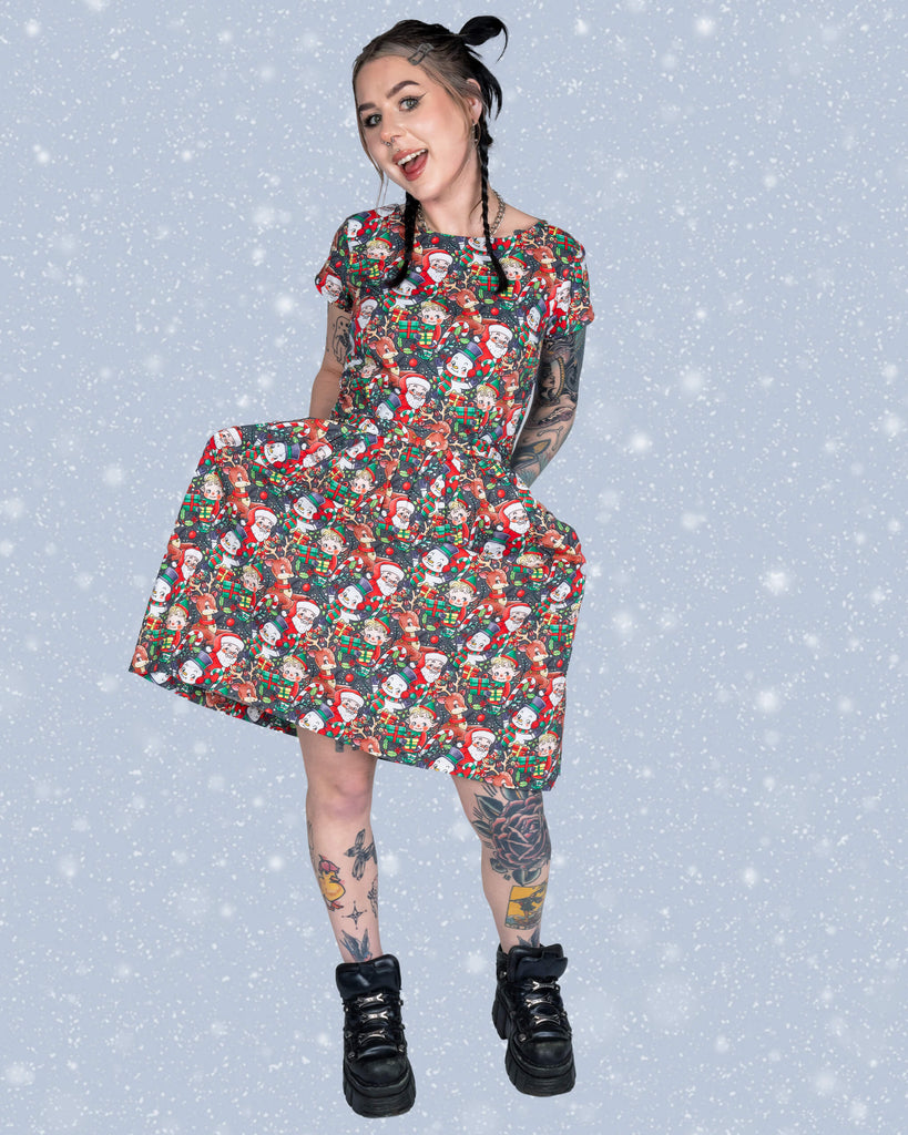 Flo grins and twirls slightly to showcase the flowy fit of the Christmas Cuties tea dress, featuring a cheerful holiday design of Santa, reindeer, and gift boxes. The snowy background adds to the festive vibe.
