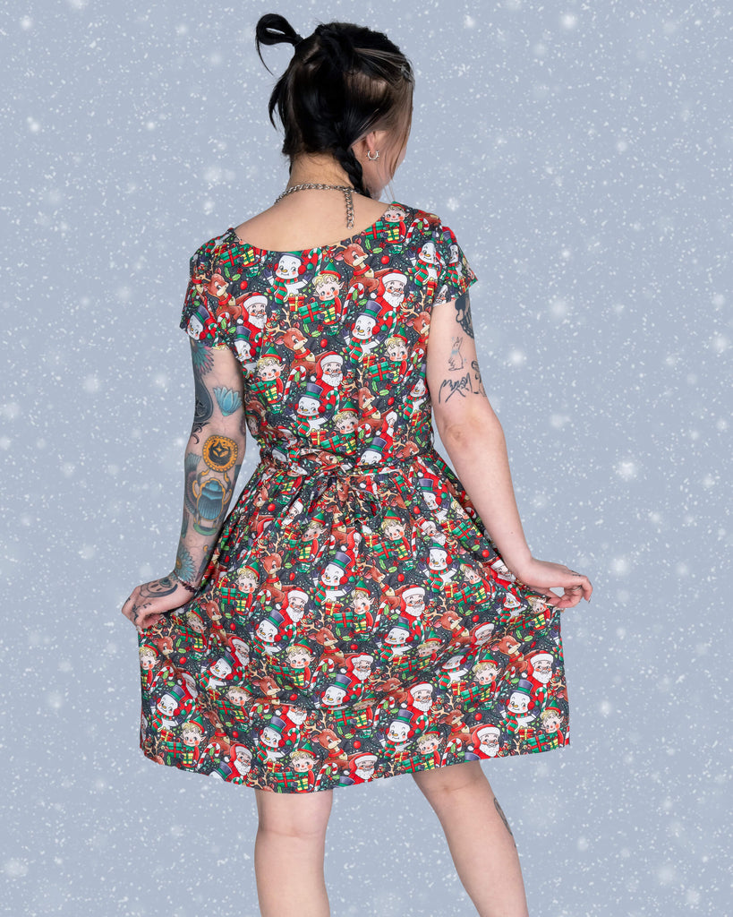 Flo turns slightly to show the back of the Christmas Cuties tea dress, highlighting its elegant shape and festive print. Her braided hair and visible tattoos contrast beautifully with the snowflake-dusted backdrop.