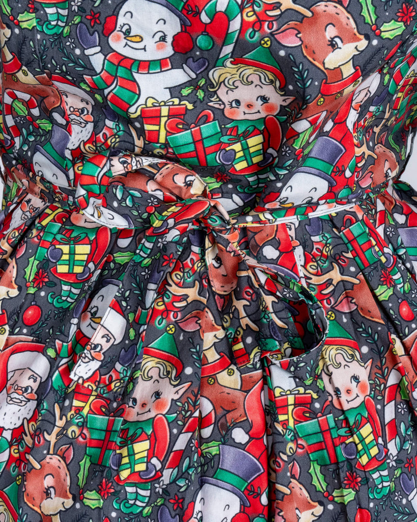 A detailed close-up of the Christmas Cuties tea dress print shows adorable holiday-themed characters, including Santa, reindeer, and elves, surrounded by gifts and holly. The dress’s pleated design and vibrant colours pop against the festive setting.