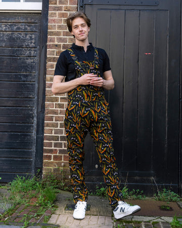 Model in Run & Fly's unisex dungarees, showcasing their quirky leaf and skull print design. The model stands casually in front of a brick and wooden background, reflecting the brand's slow fashion commitment.